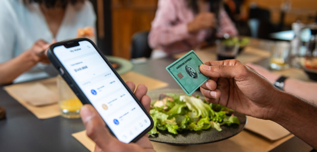 Eine Person hält eine grüne American Express Kreditkarte und ein Smartphone mit Amex App in den Händen an einem Tisch beim Mittagessen.