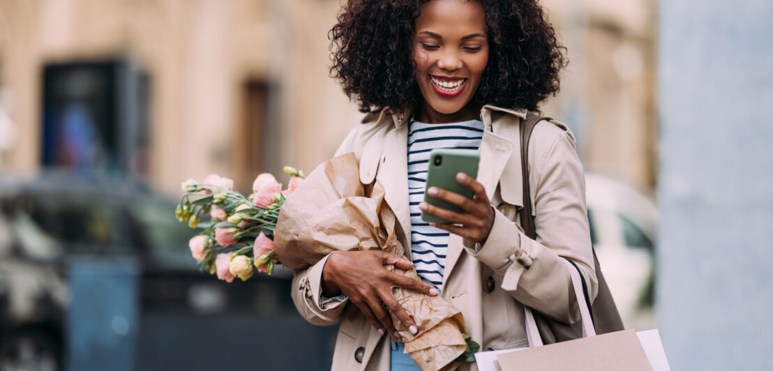 Eine Frau mit Blumenstrauß im Arm schaut lächelnd auf ihr Smartphone auf der Straße.