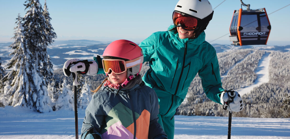 Eine Frau und ein Kind in Skianzügen auf einer Skipiste unter einer Gondel vor verschneiter Waldlandschaft.