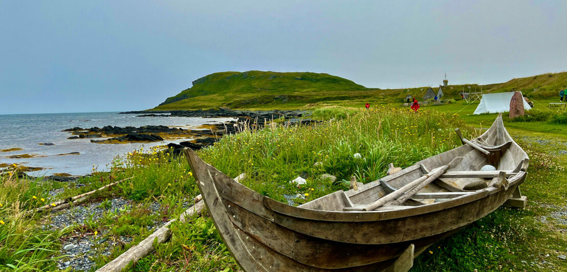 Das Bild zeigt ein altes Holzboot auf einer grünen Wiese an der Küste. Im Hintergrund sind ein Zelt und ein mit Gras bewachsenes Haus zu sehen.