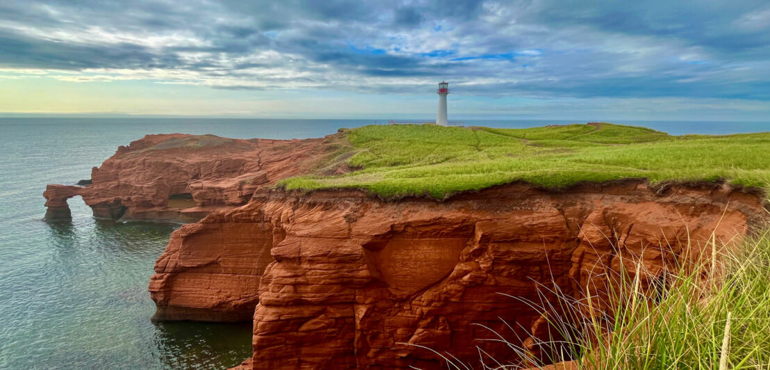 Das Bild zeigt die Steilküste im Osten von Kanada. Rote Felsen erheben sich aus dem Meer. Oberhalb sind hellgrünes Gras und ein weißer Leuchtturm zu sehen.