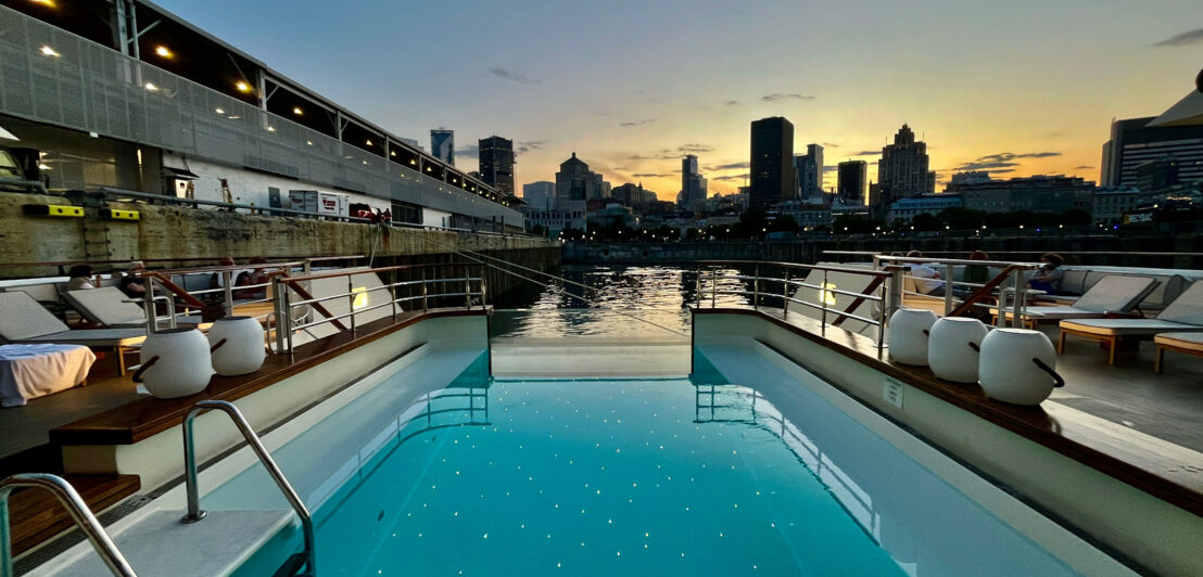 Das Bild zeigt einen Pool mit Liegen an Bord des Schiffes Le Bellot. Im Hintergrund sieht man die Umrisse der Hochhäuser der Skyline von Montreal. Dahinter geht die Sonne unter.