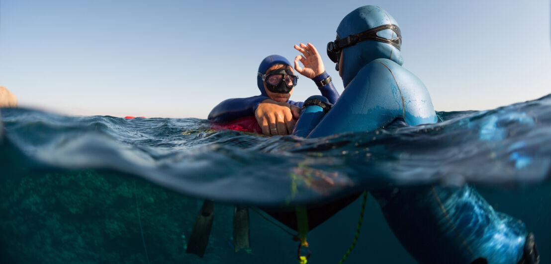 Zwei Freitaucher in Neoprenanzügen kommunizieren durch Handzeichen an einer Boje im Wasser.