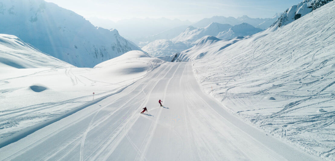 Zwei Skifahrende auf einer breiten Piste inmitten einer Schneelandschaft vor Gipfelpanorama.