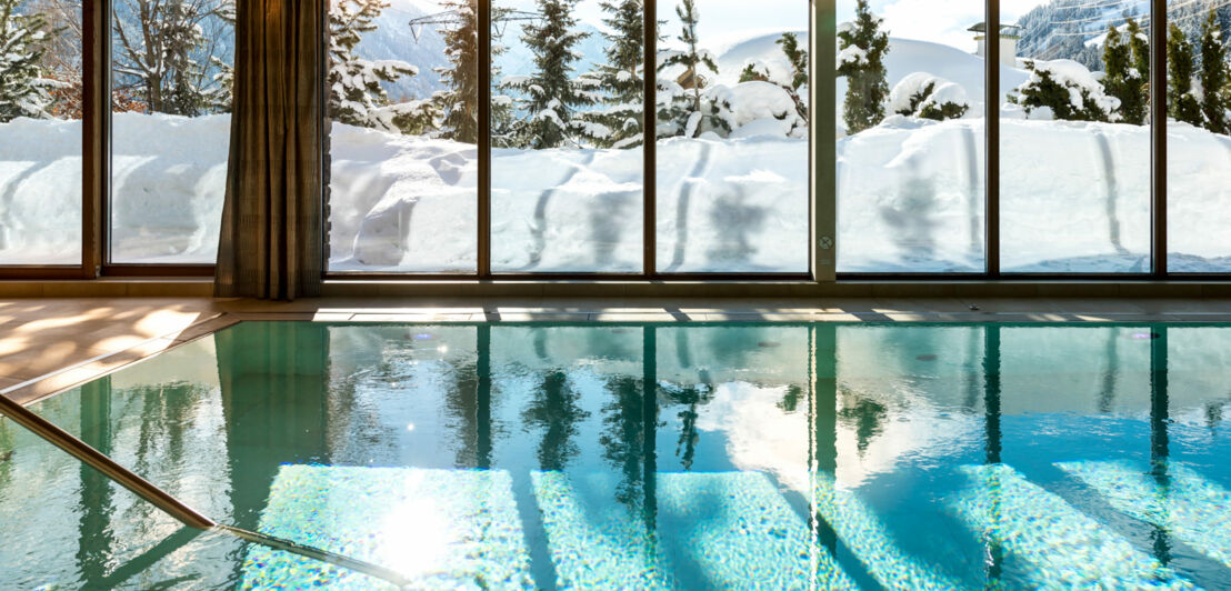 Innenpool mit Blick auf eine Schneelandschaft durch Panoramafenster bei Sonnenschein.