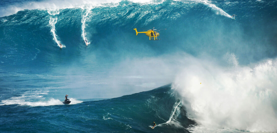 Surfende und ein Jetski auf gigantischen Wellen im Meer, über ihnen fliegt ein gelber Helikopter.