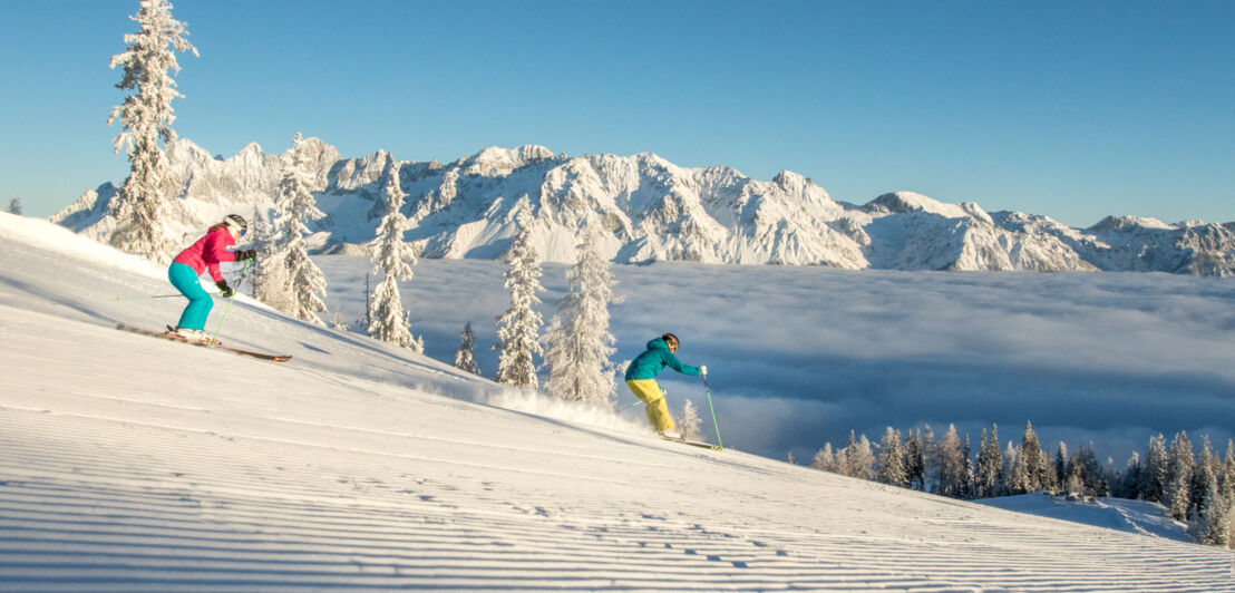 Zwei Skifahrende auf einer Piste bei der Abfahrt oberhalb der Wolkendecke vor schneebedeckten Gipfeln.