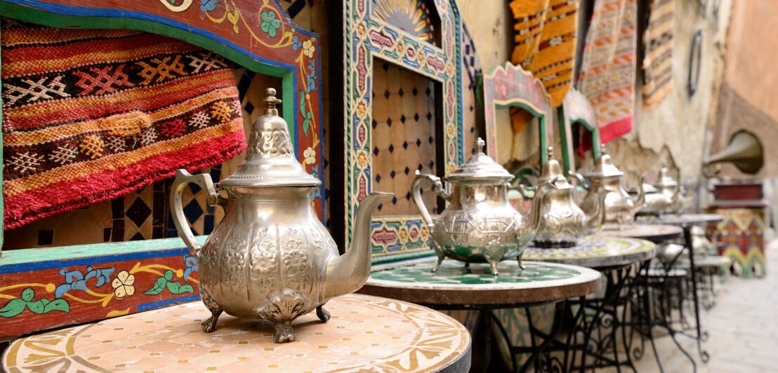 Orientalische Teekannen auf runden Mosaiktischen vor einer Wand mit Berberteppichen.