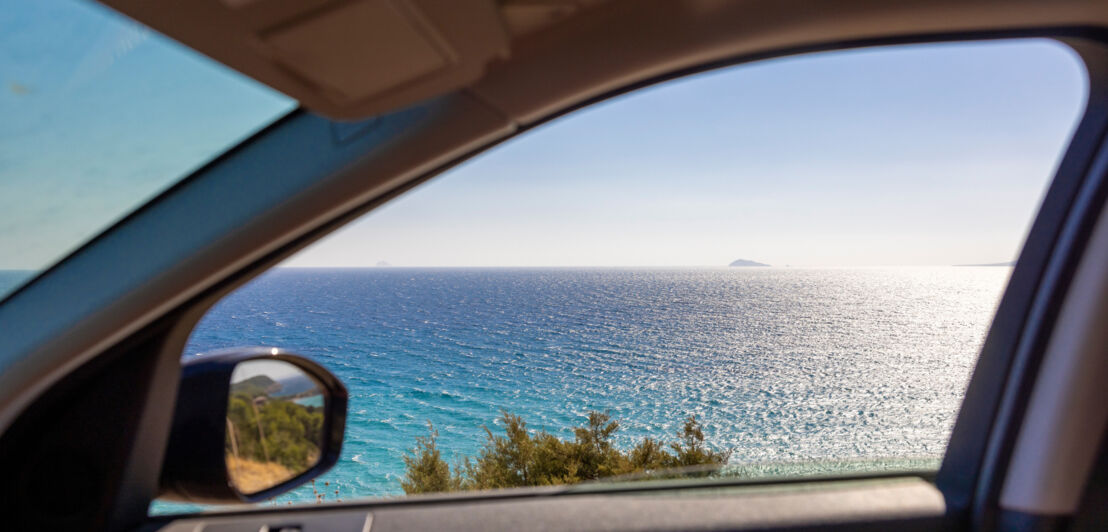 Meerblick durch ein geöffnetes Autofenster an einer Küste bei Sonnenschein.