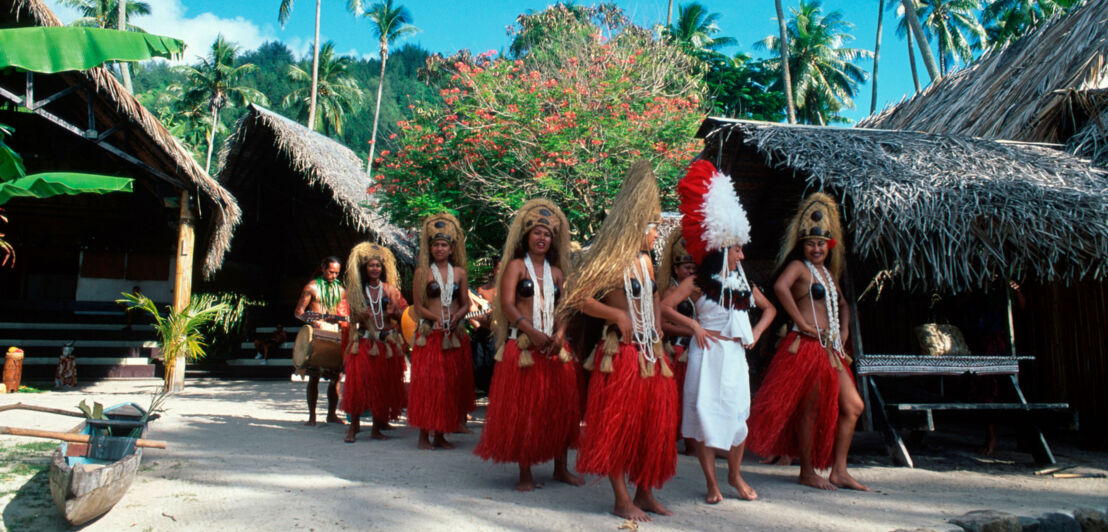 Aufgereihte Frauen mit roten Baströcken und Kopfschmuck in einem polynesischen Dorf.