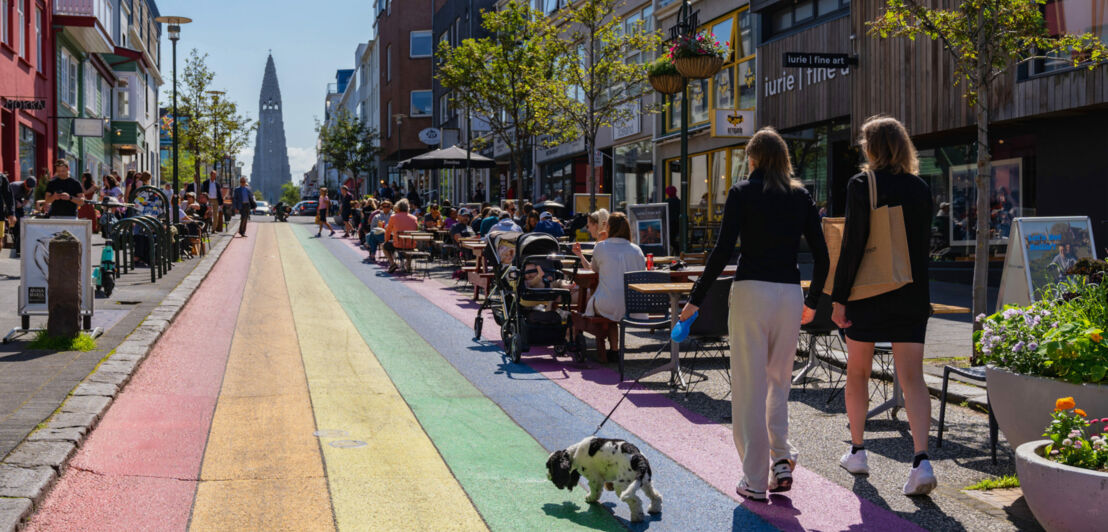 Personen auf einer Einkaufsstraße mit Regenbogenfarben auf dem Asphalt bei Sonnenschein.