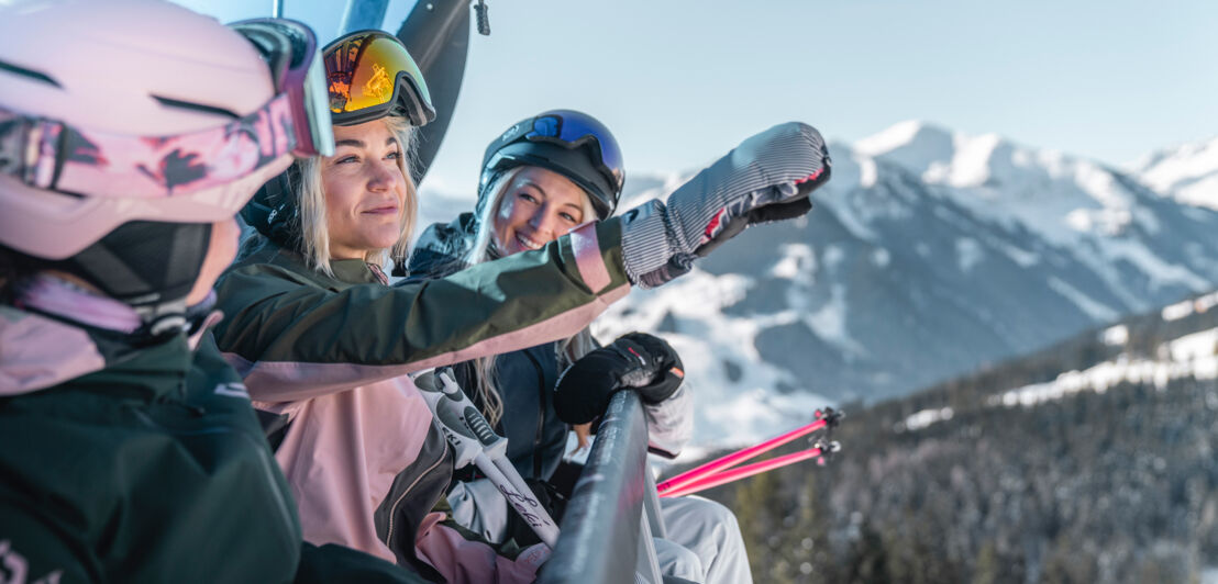 Drei Skifahrerinnen machen ein Selfie in einem Sessellift vor schneebedecktem Bergpanorama.