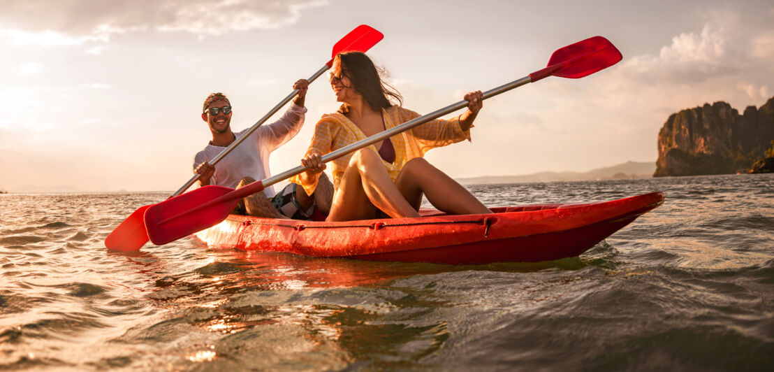 Zwei glückliche Personen, die bei Sonnenuntergang in einem Kajak auf dem Wasser sitzen und jeweils ein Padel halten.