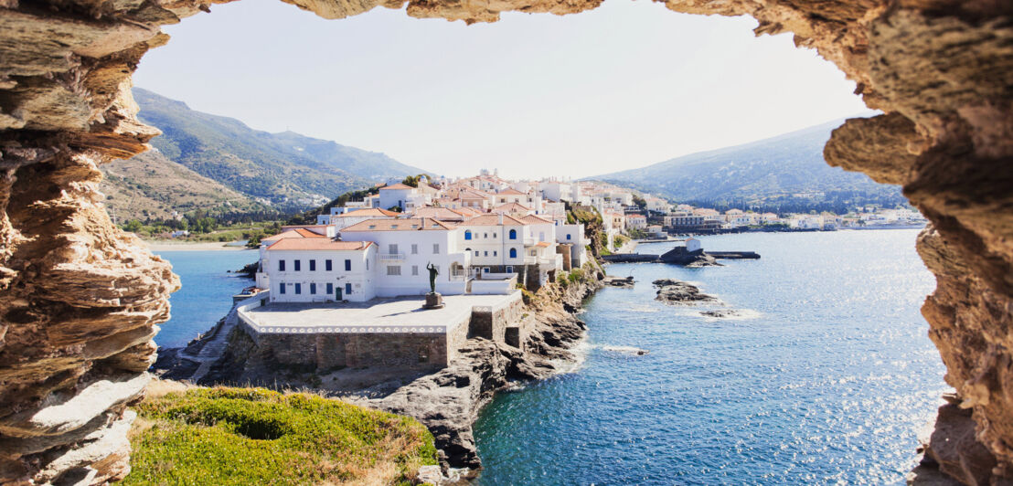 Blick auf eine Landzunge mit weißen Häusern an einer mediterranen Küste durch ein Loch in einem Felsblock.