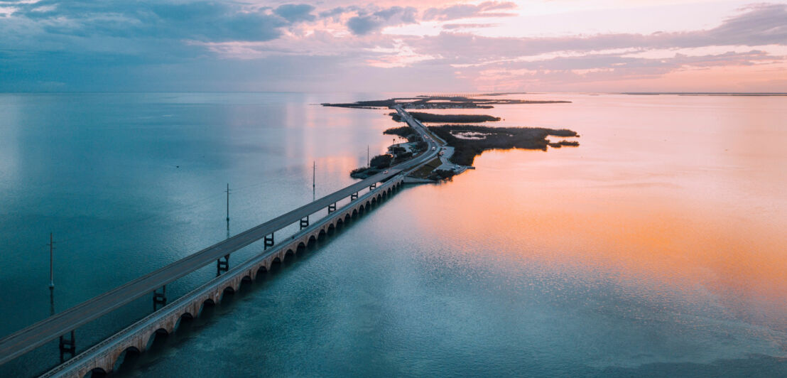 Eine lange, zweispurige Autobrücke über Wasser, die kleine Inseln miteinander verbindet bei Sonnenuntergang.