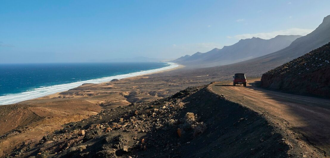 Ein Jeep fährt auf einer unbefestigten durch eine gebirgige Wüstenlandschaft am Meer.