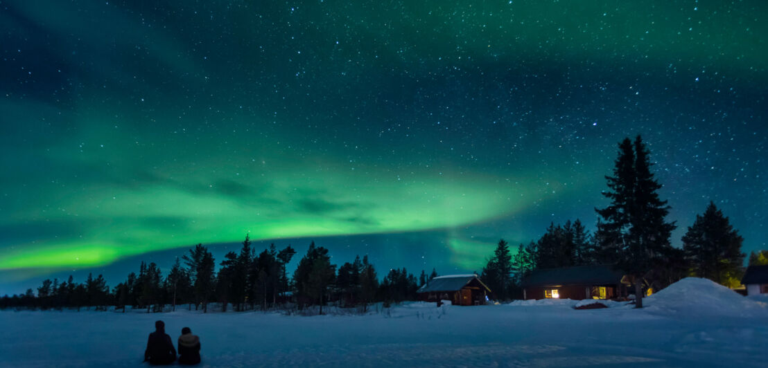 Zwei Personen sitzen im Schnee unter einem Sternenhimmel mit Polarlichtern.