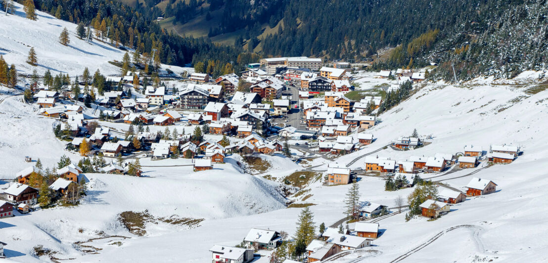 Schneebedeckter Ort in einem Tal, umgeben von bewaldeten Bergen.