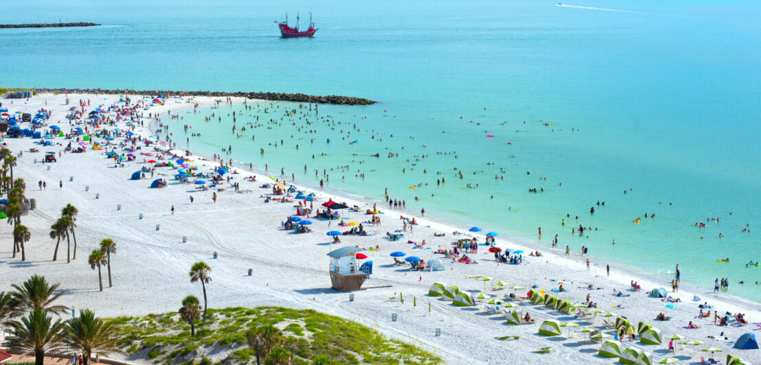 Belebter, breiter Strand mit weißem Sand an türkisblauem Wasser.