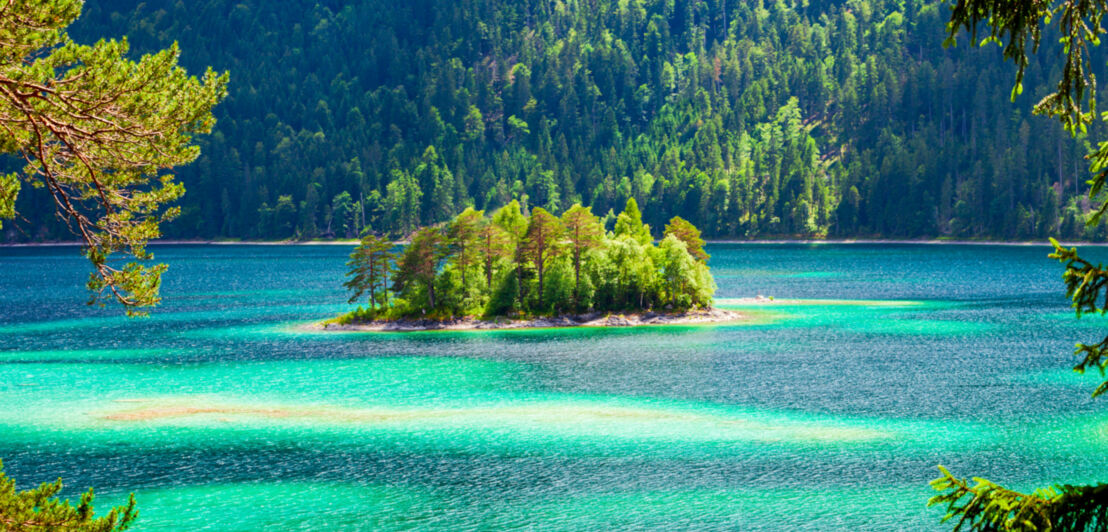 Ein See in verschiedenen Türkis- und Blautönen, in der Mitte eine kleine Insel und im Hintergrund ein Wald.