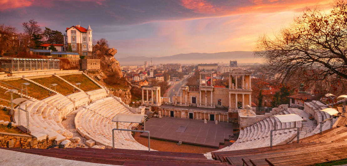 Antikes Amphitheater vor Stadtpanorama bei Sonnenuntergang.