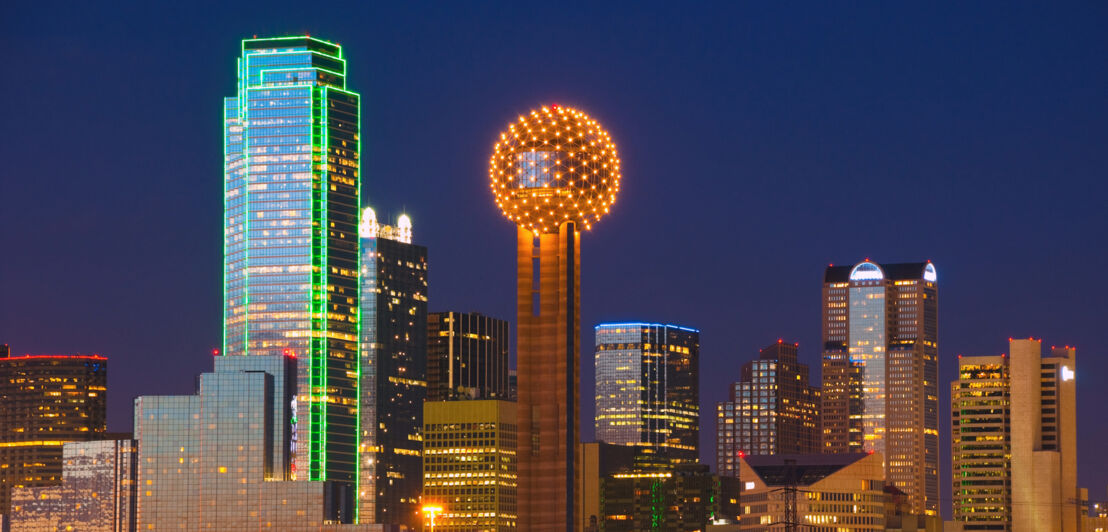 Erleuchtete Skyline von Dallas mit Wolkenkratzern und Gebäude mit kugelförmiger Spitze bei Dunkelheit.