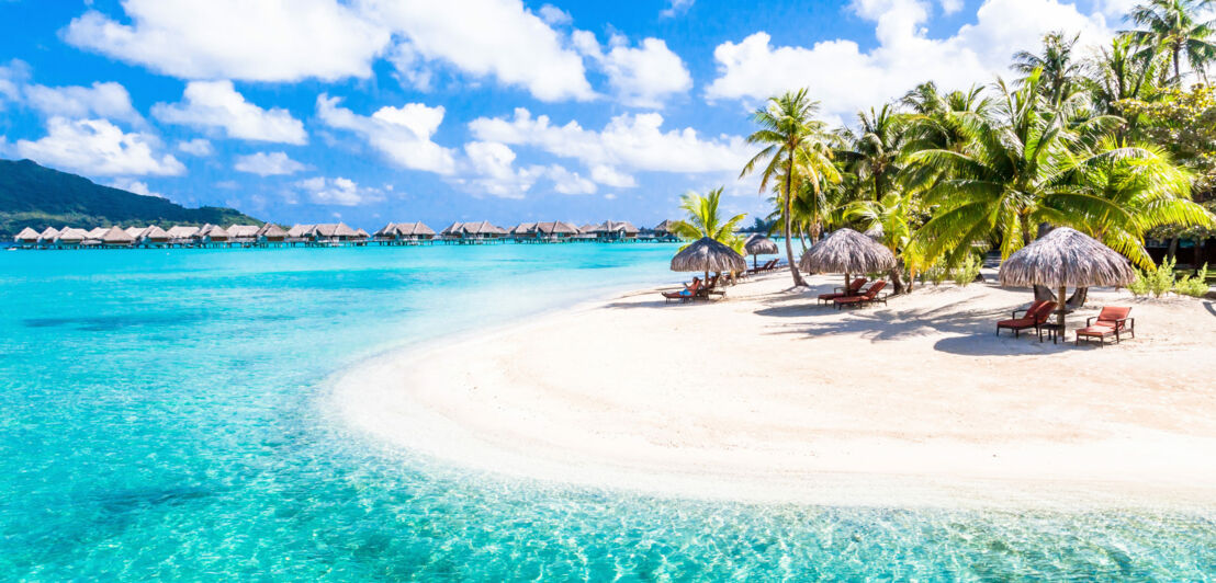 Strandliegen mit Strohschirmen auf einem palmengesäumten Strand mit weißem Sand an türkisblauem Wasser, im Hintergrund Hotelresort mit Überwasser-Bungalows.