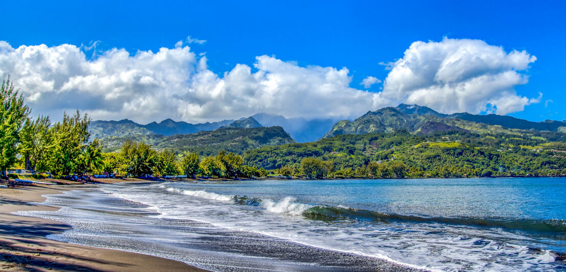 Menschenleerer Strand mit grüner Vegetation vor Berglandschaft.