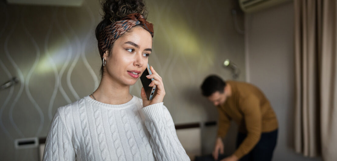 Eine Frau, die ein Smartphone an ihr Ohr hält, im Hintergrund steht ein Mann über einen Koffer gebeugt.