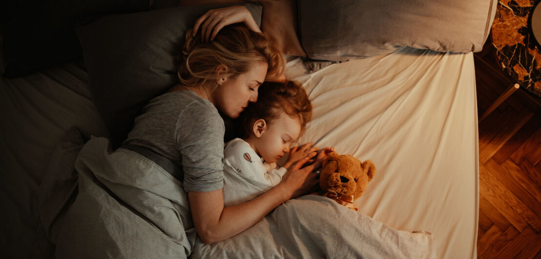 Eine Frau und ein Kind liegen aneinandergekuschelt mit einem Teddy im Bett.