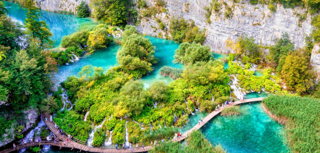 Blick von oben auf einen türkisblauen See, durch den ein Holzsteg führt und der von Bäumen und Felsen umgeben ist.