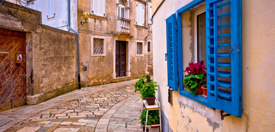 Eine schmale Altstadtgasse, im Vordergrund ein Haus mit blauen Fensterläden und Blumen vor den Fenstern.