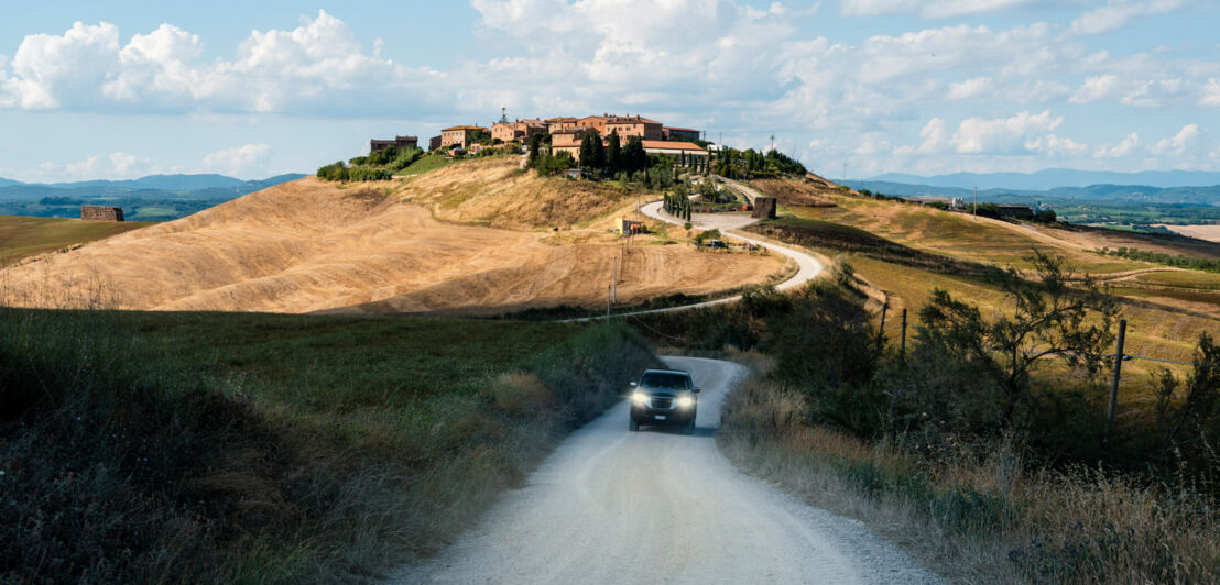 Ein SUV auf einer unbefestigten, kurvigen Straße in mediterraner Landschaft, im Hintergrund eine Ortschaft auf einem Hügel.