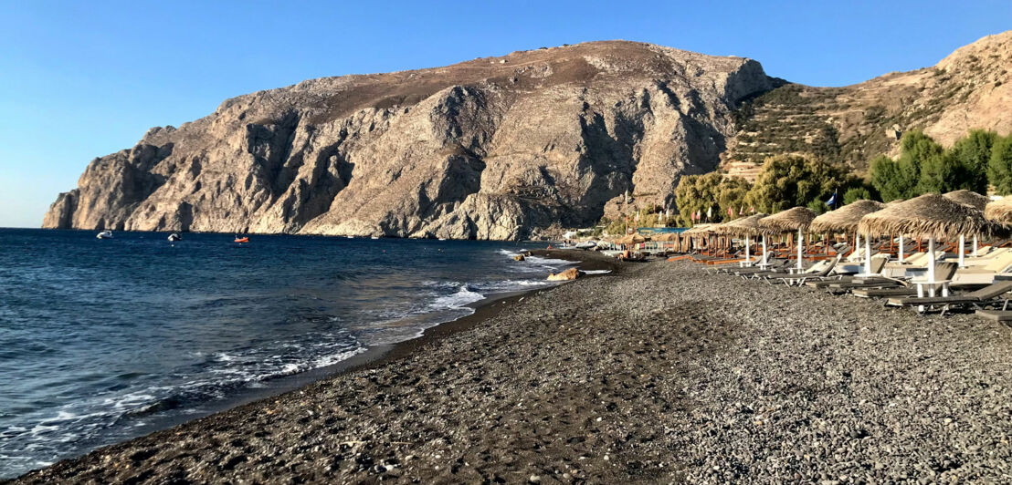 Schwarzer Sandstrand mit Sonnenliegen an einer Felsküste.