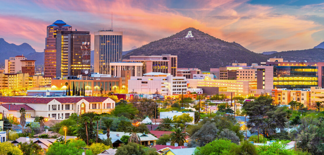 Beleuchte Skyline von Tucson mit Hochhäusern und grüner Vegetation vor Gebirgszug bei Sonnenuntergang.