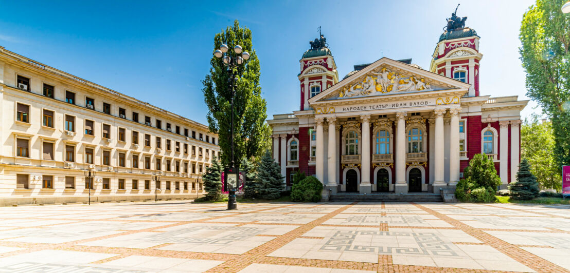 Historisches Theatergebäude mit Dreiecksgiebel und Säulen am Eingangsbereich an einem Platz.