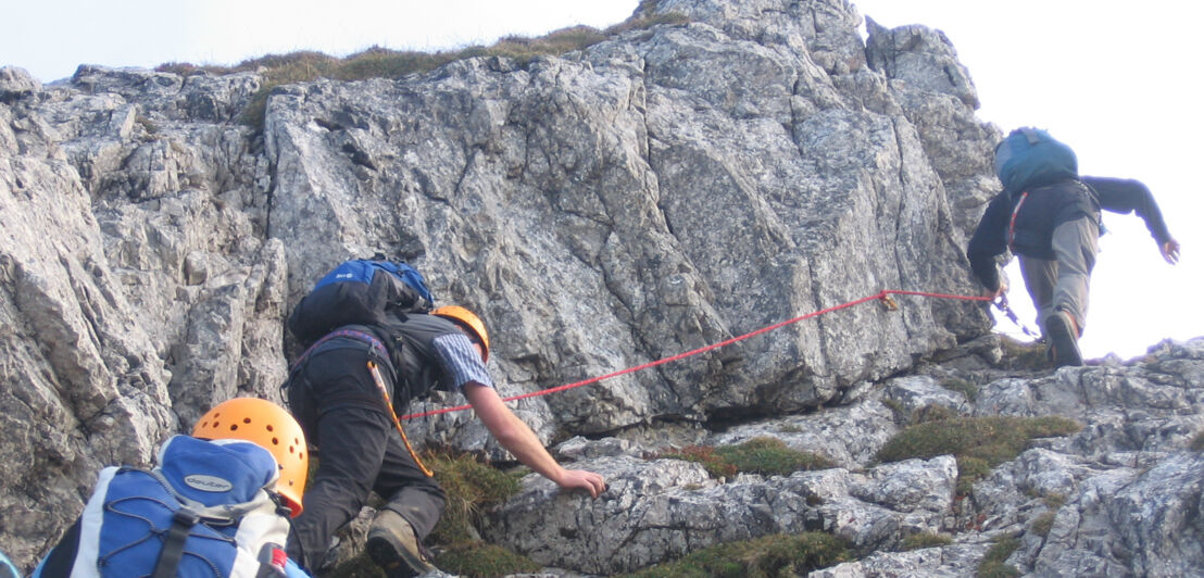 Drei Personen mit Kletterausrüstung, die einen steilen Felsen hochklettern, an dem ein Seil gespannt ist. Drei Personen mit Kletterausrüstung, die einen steilen Felsen hochklettern, an dem ein Seil gespannt ist.