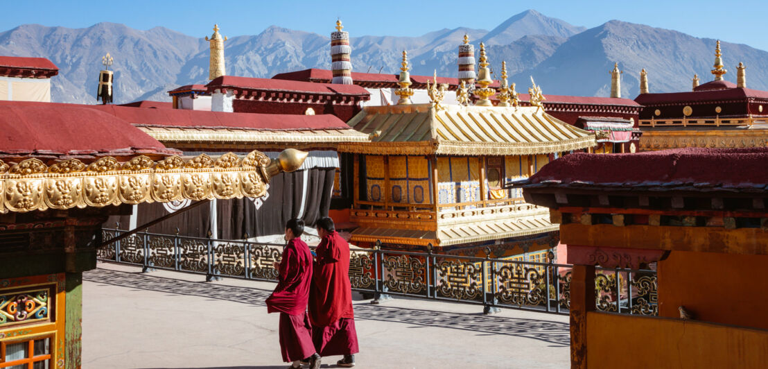 Zwei tibetische Mönche in roten Kutten gehen durch eine buddhistische Tempelanlage vor Bergpanorama.