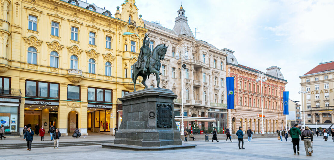 Personen auf einem historischen Platz mit Häusern in unterschiedlichen Architekturstilen und Reiterstandbild.