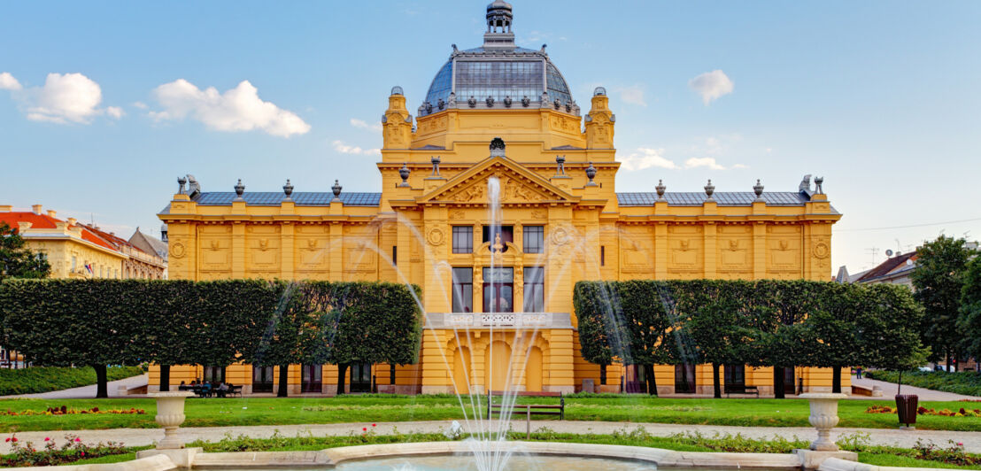 Gelbes Jugendstilgebäude mit Glaskuppel in einer Parkanlage mit Springbrunnen im Vordergrund.