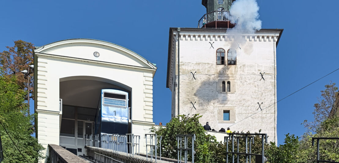 Weißer Turm mit Dampf auf dem Dach aus einer Kanone neben einer Standseilbahnstation auf einem Hügel.