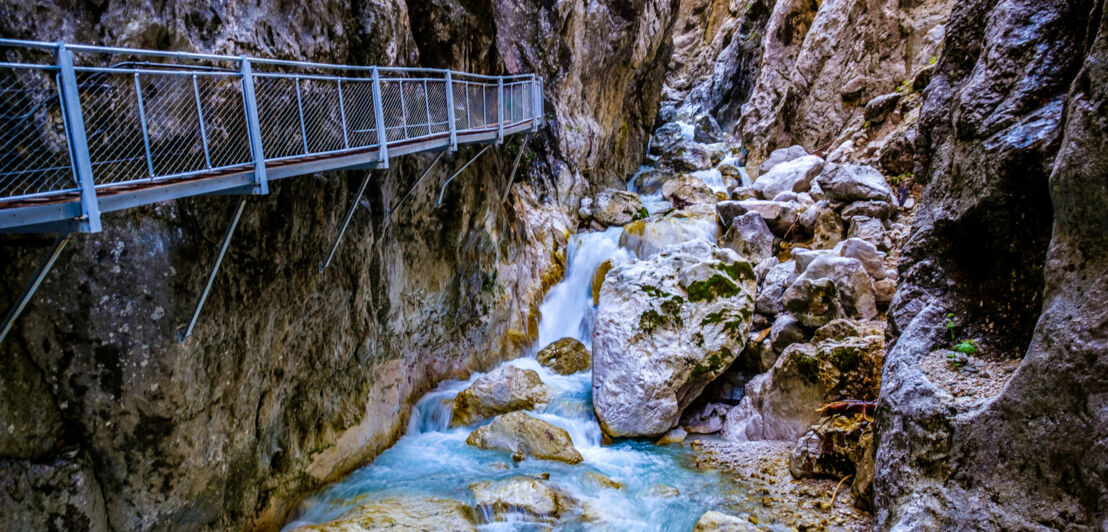 Enge Schlucht mit einem kleinen Wasserfall und einer schmalen Brücke an einer Felswand. 