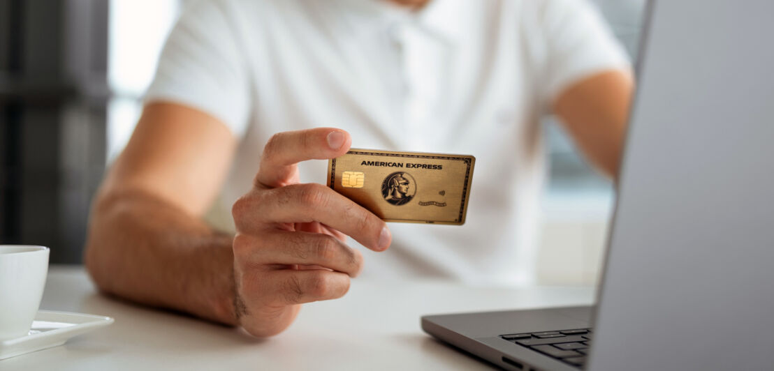 Nahaufnahme einer goldenen American Express Kreditkarte in der Hand einer Person bei der Online-Zahlung an einem Laptop.