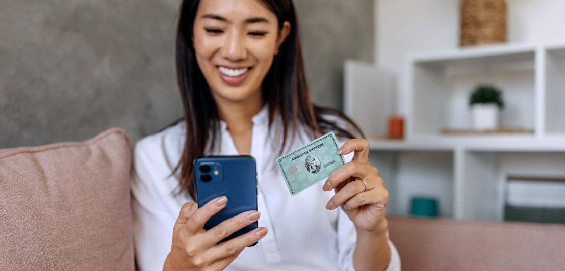 Eine Frau sitzt auf einem Sofa und blickt lächelnd auf ein Smartphone in ihrer Hand, in der anderen Hand hält sie eine grüne American Express Kreditkarte.