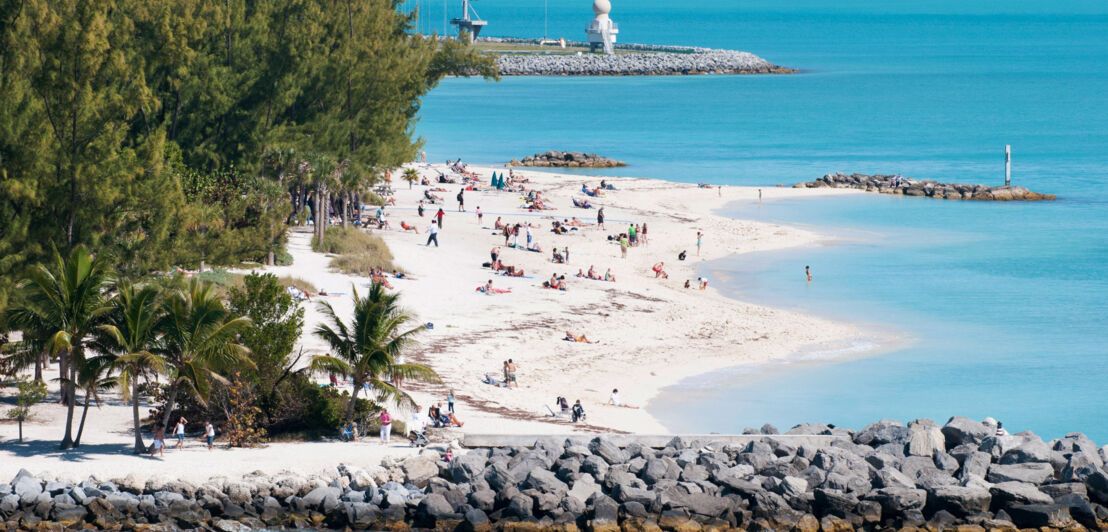 Personen an einem weißen Sandstrand mit Bäumen am türkisblauen Meer, im Vordergrund ein Wellenbrecher aus aufgeschütteten Steinen.