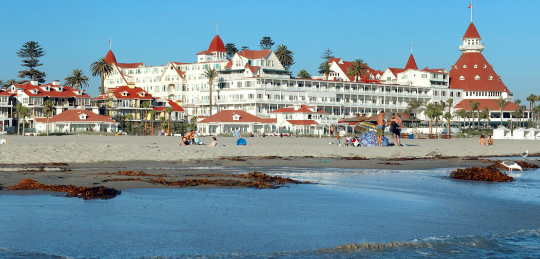 Blick vom Wasser auf einen Sandstrand mit Palmen und Personen vor einem weißen Hotelkomplex im viktorianischen Stil.