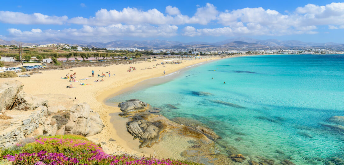 Personen auf einem hellen Sandstrand am türkisblauen Meer in mediterraner Umgebung.