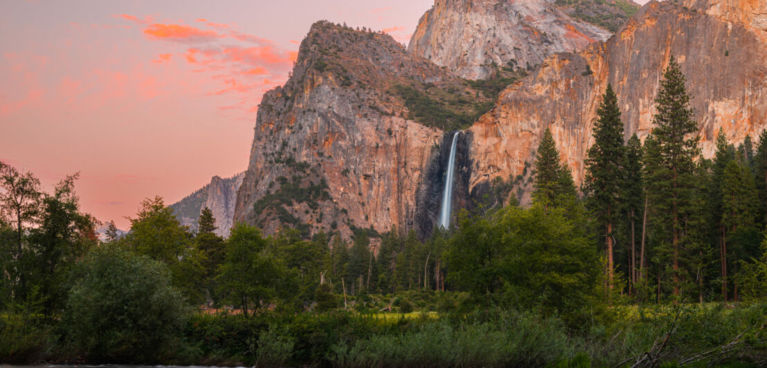 Ein Wasserfall an einer Bergwand, im Vordergrund ein See in einem Waldgebiet im rötlichen Abendlicht.