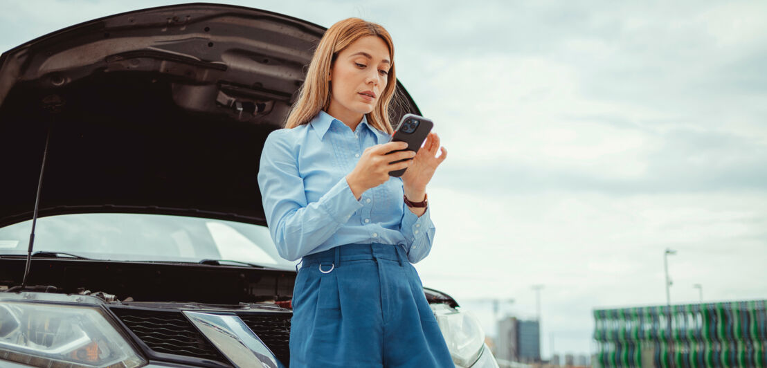 Eine Frau lehnt an einem Auto mit geöffneter Kühlerhaube und bedient ihr Smartphone.