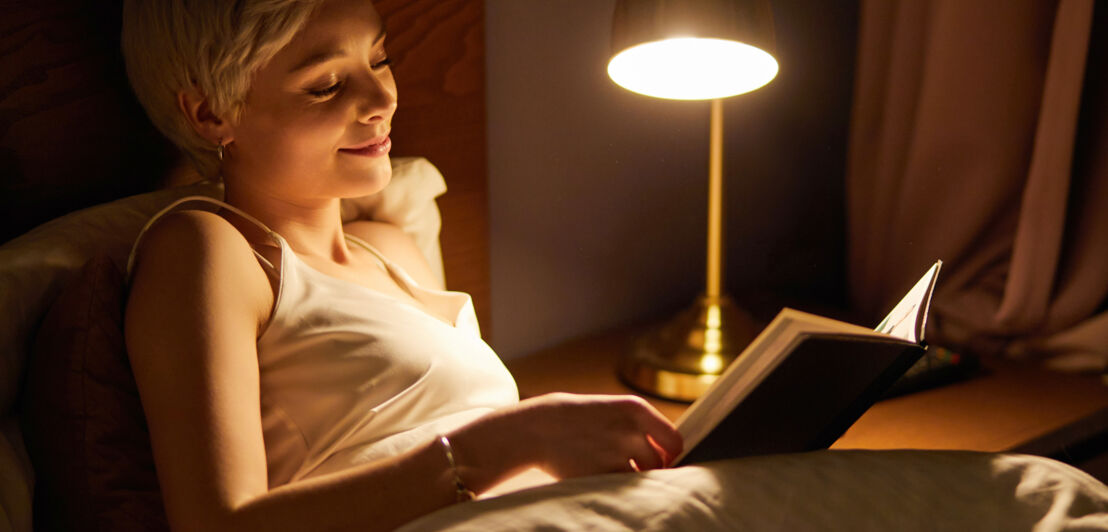 Eine Frau liest im Bett ein Buch im Licht einer Nachttischlampe.